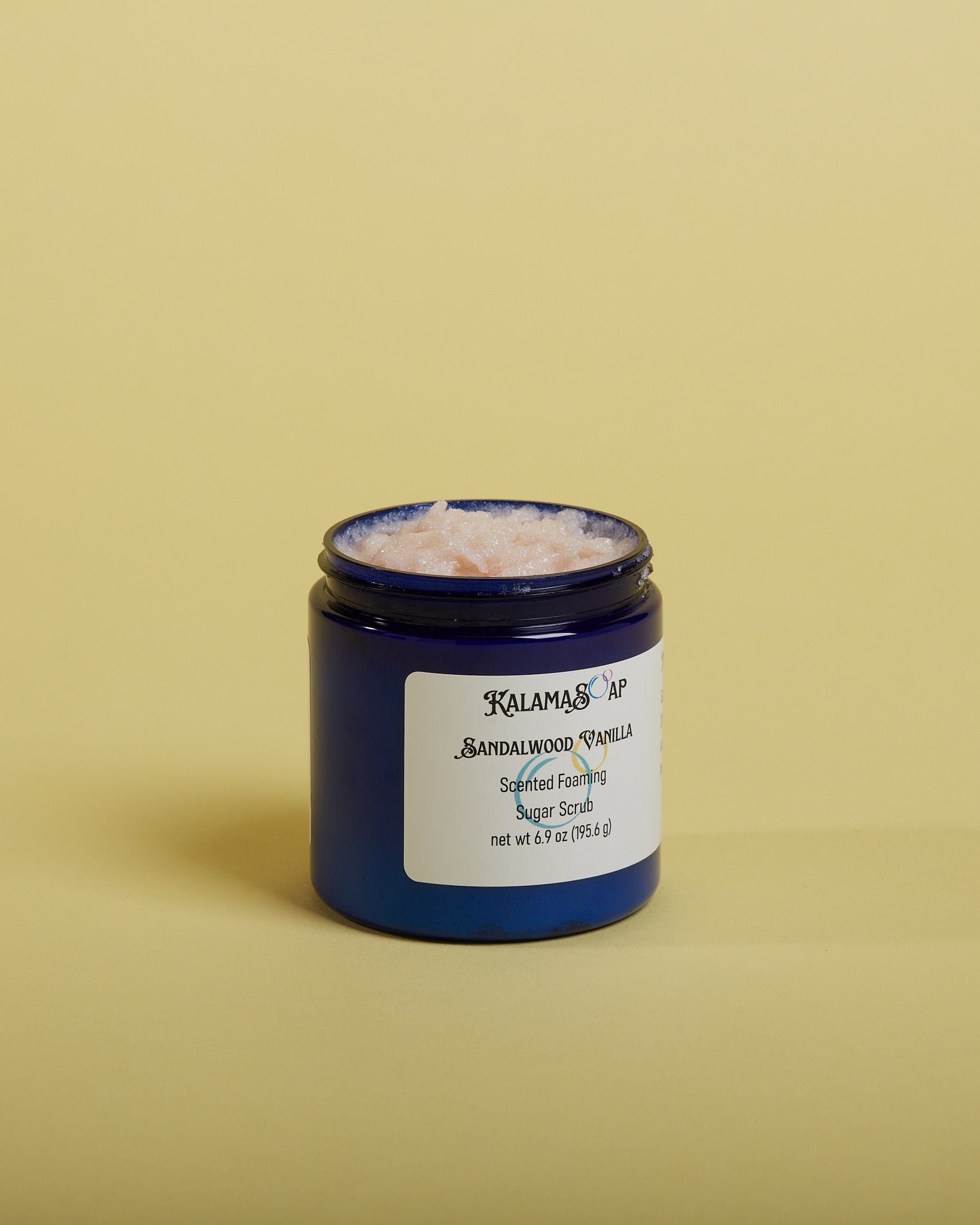 A photo of an open 6.9 ounce blue plastic jar displaying the sugar scrub contents on a yellow background labeled KalamaSoap Sandalwood Vanilla Scented Foaming Sugar Scrub