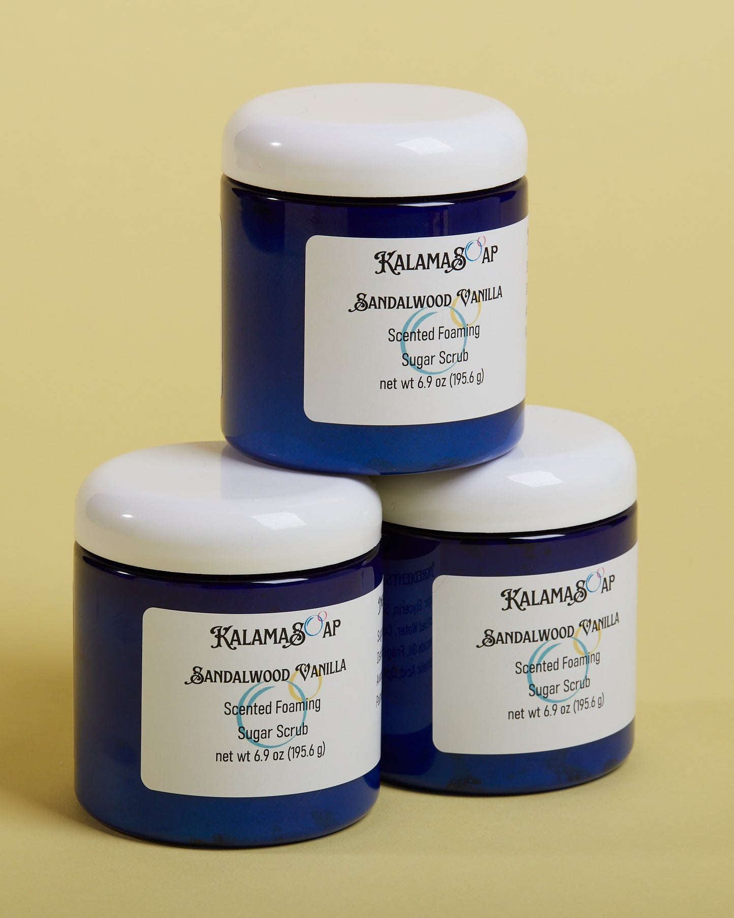 A photo of 3, 6.9 ounce blue plastic jars with white dome lids on a yellow background labeled KalamaSoap Sandalwood Vanilla Scented Foaming Sugar Scrub