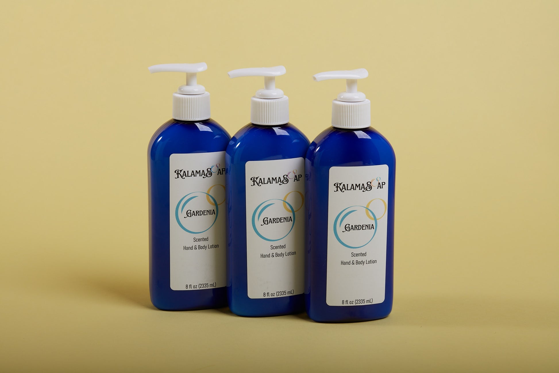 A photo of 3, 8-fluid ounce blue lotion bottles with white pumps on a yellow background labelend KalamaSoap Gardenia Scented Hand & Body Lotion