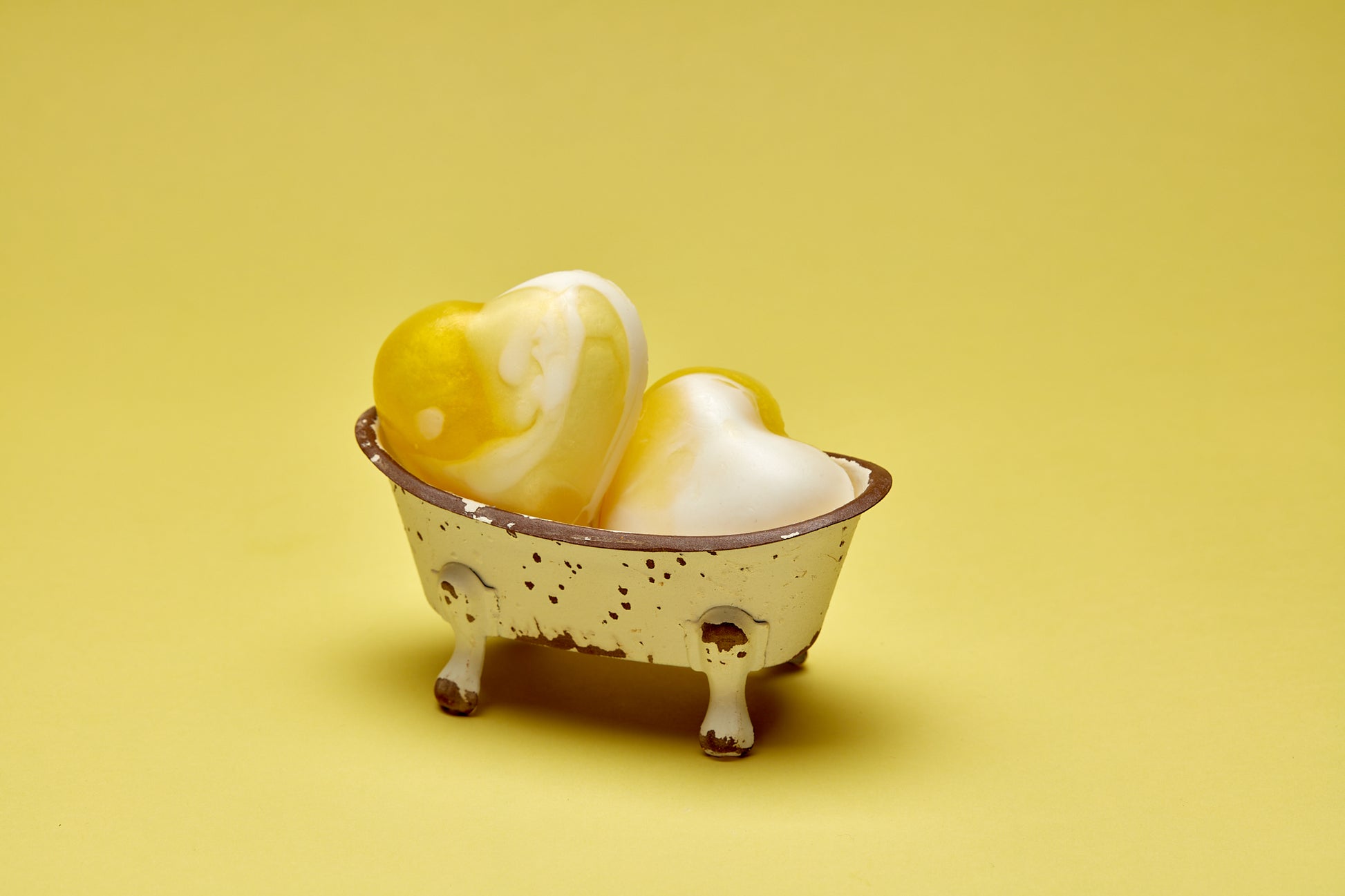 A photo of 2 heart shaped yellow and white Gardenia scented soaps in a distressed mini bath tub on a yellow background.