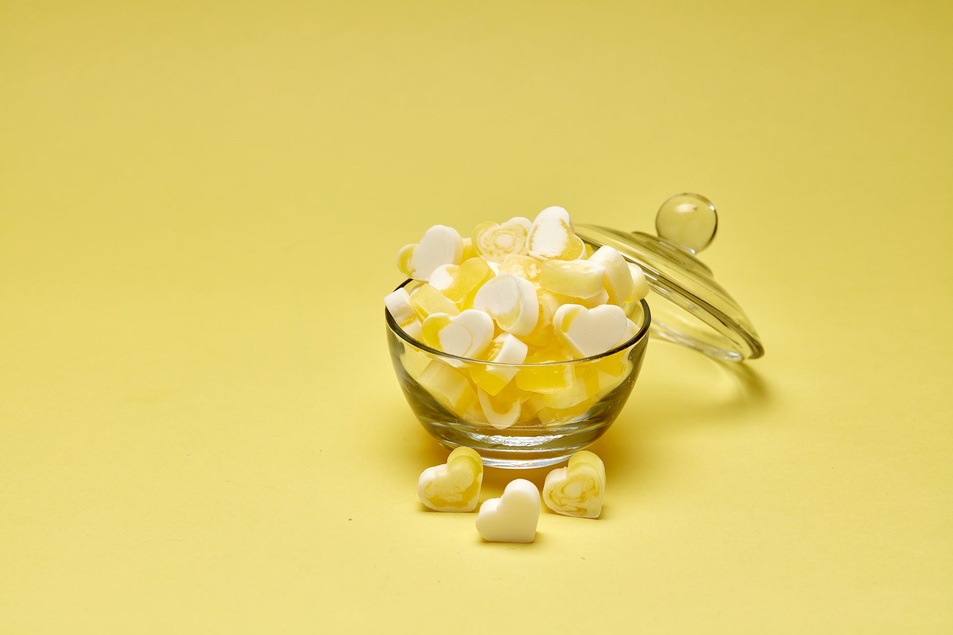 A photo of heart shaped yellow and white mini soaps in a glass bowl, lid leaning against it on a yellow background.