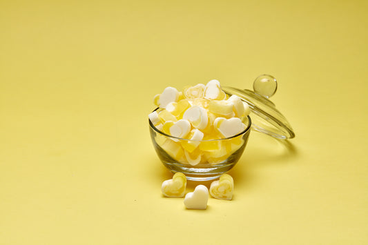 A photo of heart shaped yellow and white mini soaps in a glass bowl, lid leaning against it on a yellow background.