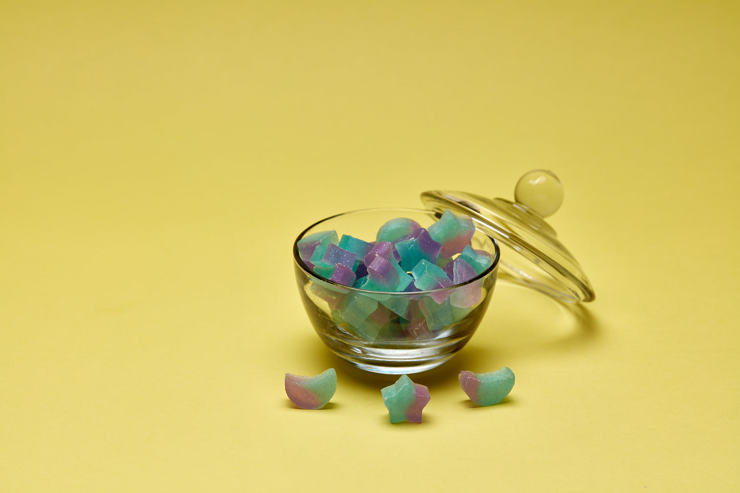A photo of moon and star shaped blue/green and purple with glitter mini soaps in a glass bowl, lid leaning against it on a yellow background.