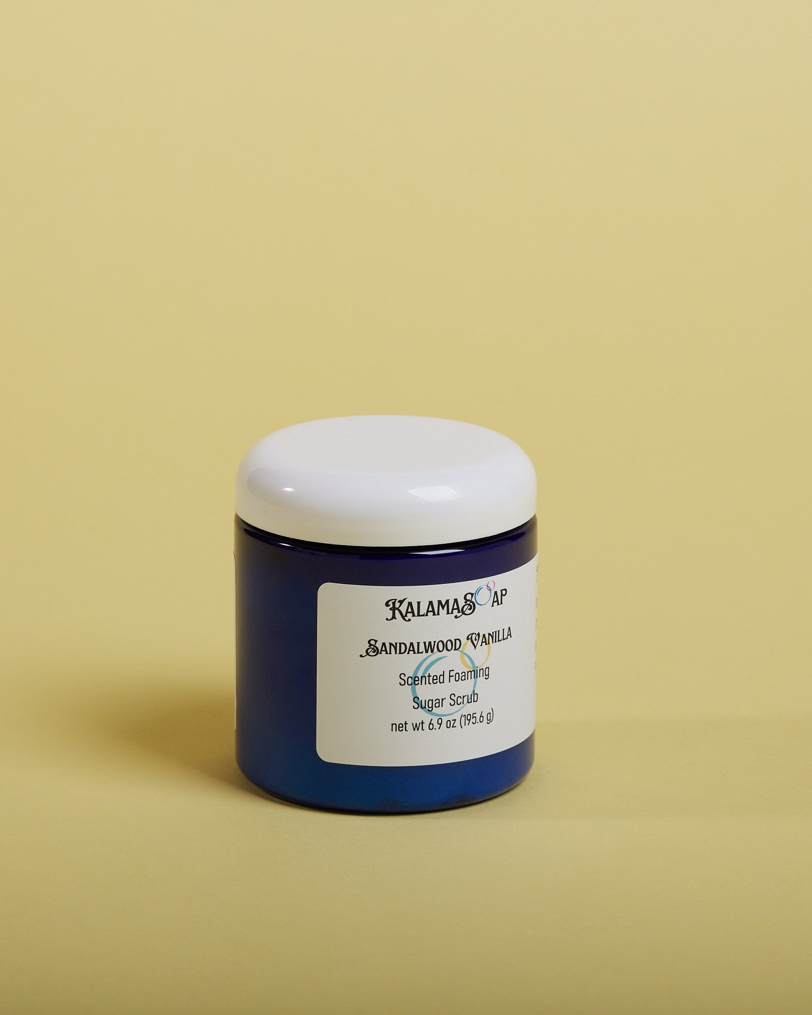 A photo of a 6.9 ounce blue plastic jar with white dome lids on a yellow background labeled KalamaSoap Sandalwood Vanilla Scented Foaming Sugar Scrub