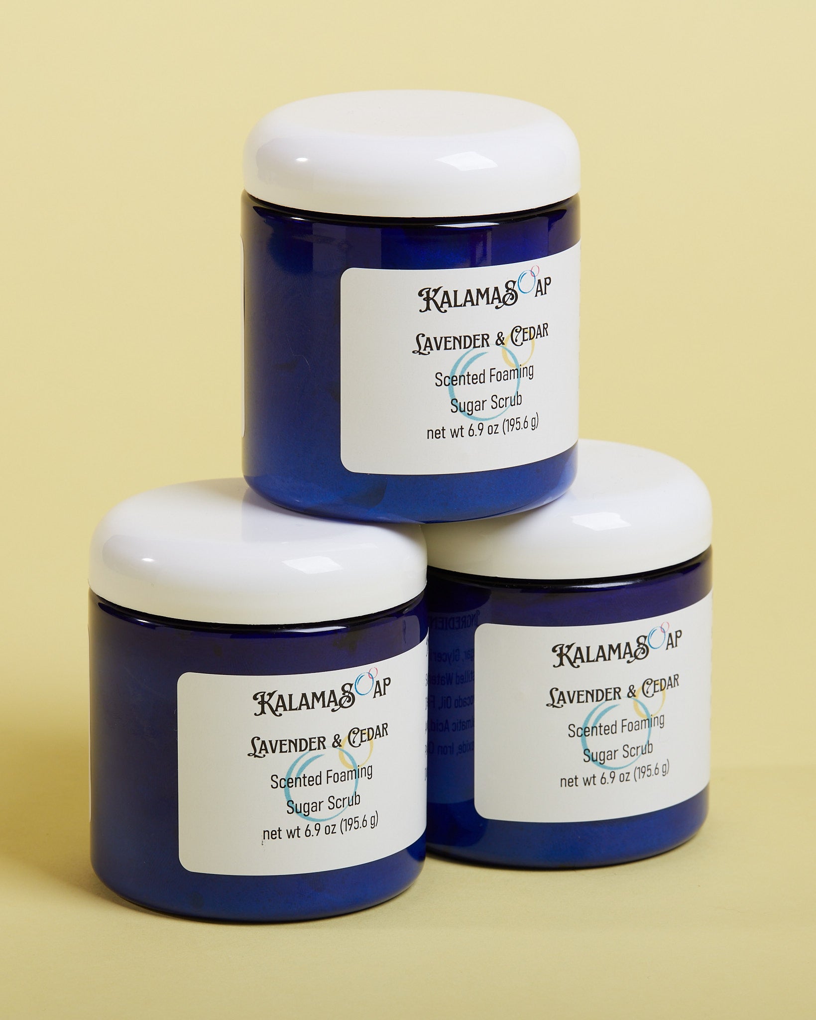A photo of 3, 6.9 ounce blue plastic jars with white dome lids on a yellow background labelend KalamaSoap Lavender & Cedar Scented Foaming Sugar Scrub
